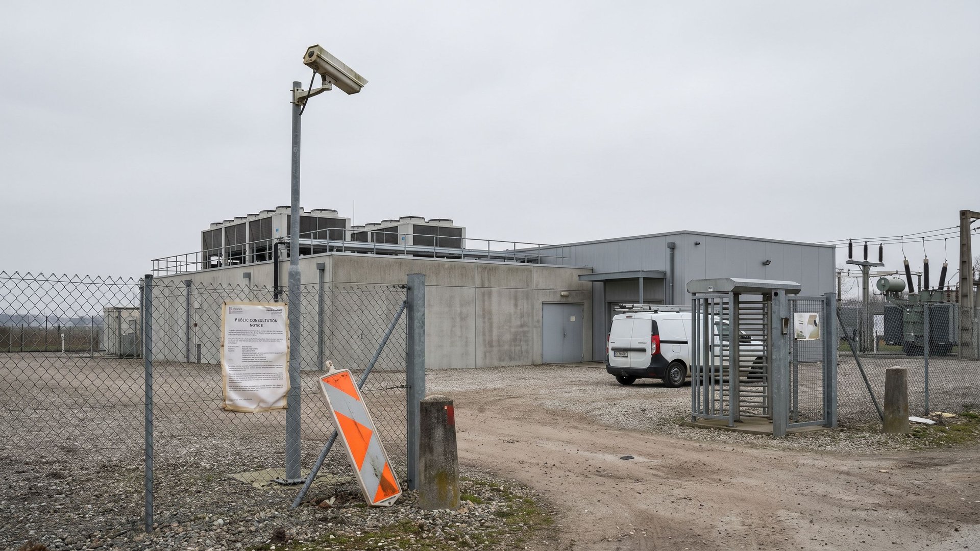 Low-lying data center complex behind a chain-link perimeter fence with rooftop cooling units and a laminated public consultation notice stapled to the gate